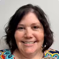 A woman with wavy dark hair and light skin smiles at the camera. She is wearing a colorful, patterned top and is pictured against a plain, light-colored background.